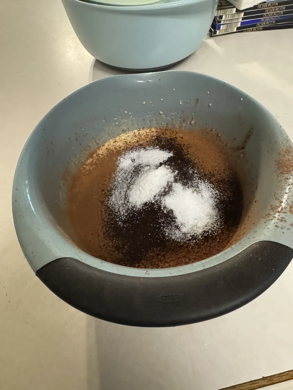 a mixing bowl containing brown batter, with various white and dark powders resting on the surface
